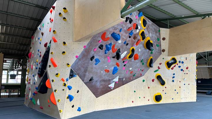Neu und glänzende Boulder an einer Boulderwand mit Griffen in verschiedene Farben. Die Farbe der Wände ist teil hellbraun, teils dunkelbraun. Unterhalb der Wände liegt einen Fallschutzmatte in dunkelgrauer Farbe