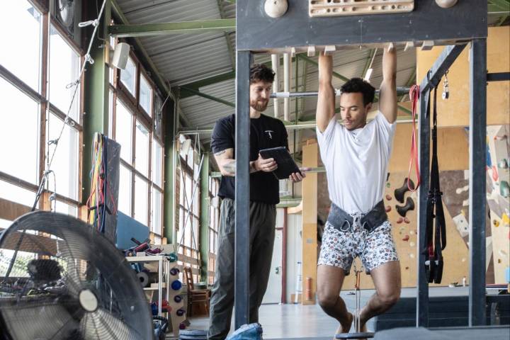 Ein Mann hängt mit beiden Armen an einem Holzbrett. Er wird angeleitet von einem weiteren Mann, welcher neben ihm auf dem Boden steht. Dieser Mann ist der Trainer beim steilen Training.