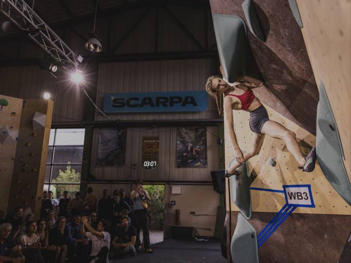 Eine junge blonde Frau ist in einem Finalboulder des ESC Finale in der Boulderhalle Steil in Karlsruhe  zu sehen. Im Hintergrund sind die Zuschauerränge voll mit Menschen.